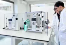 FABRX M3DIMAKER Enables Scalable Automated Capsule Filling Two scientists in white lab coats examine a glass-enclosed laboratory device on a clean white bench behind them are other equipment pieces and glass walls.