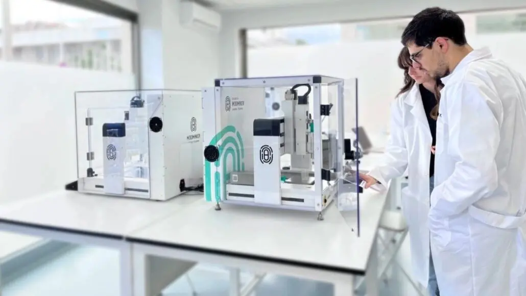 Two scientists in white lab coats examine a glass-enclosed laboratory device on a clean white bench behind them are other equipment pieces and glass walls.