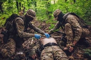 Two soldiers in camouflage treat a wounded person lying on the forest floor with first aid.