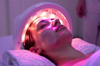 Person lies with eyes closed as a red LED facial device treats their face with a pink glow in a spa setting.