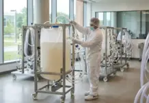 Single Use Facility Design Supporting Biopharma Agility Healthcare worker in full PPE adjusts a large IV bag on a wheeled medical cart in a clean laboratory setting with large windows.