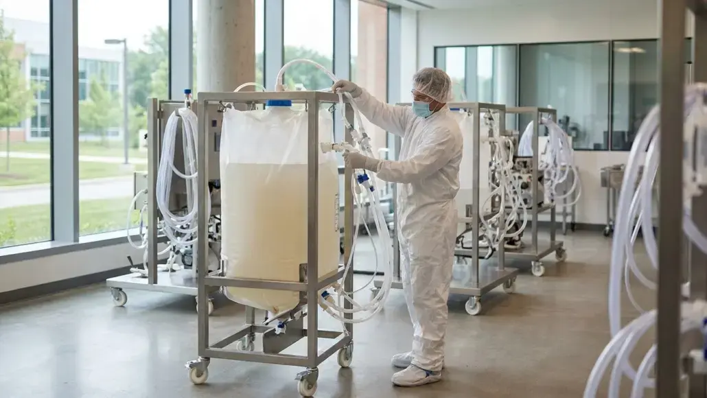 Healthcare worker in full PPE adjusts a large IV bag on a wheeled medical cart in a clean laboratory setting with large windows.