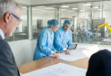Annex 1 Compliance Reshaping Sterile Facility Design Three engineers in blue lab coats and hairnets review blueprints on a table in a cleanroom setting.