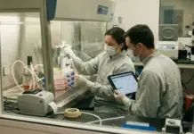 Breaking the 14-Day Barrier with Modern CAR-T Manufacturing Two lab technicians in masks inside a biosafety cabinet pipette liquids into stacked tubes while one uses a tablet to record data in the experiment workflow.